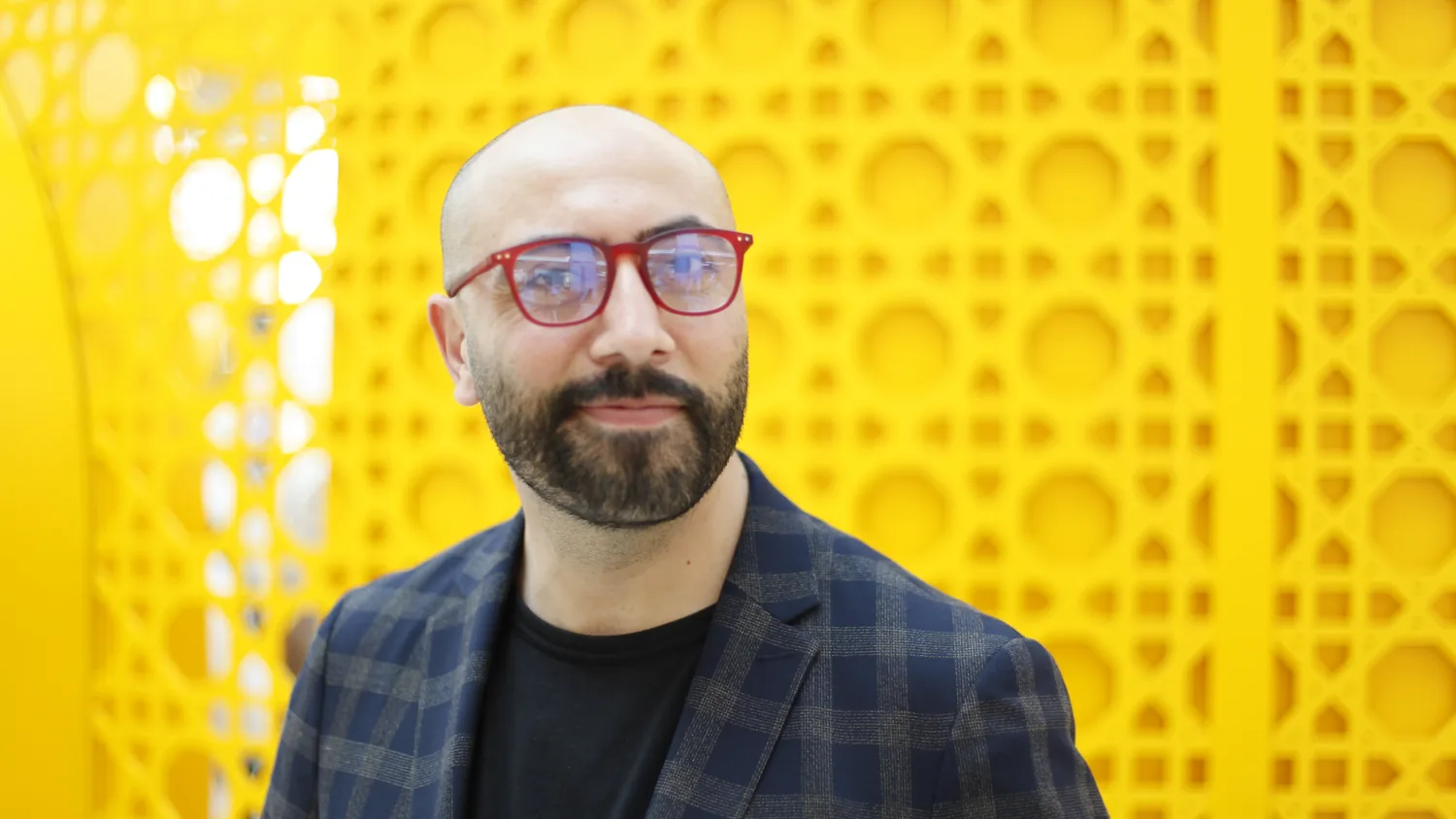 Ali Al Mokdad in red glasses and a checked jacket, photographed against a bright yellow perforated wall.