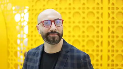 Ali Al Mokdad in red glasses and a checked jacket, photographed against a bright yellow perforated wall.