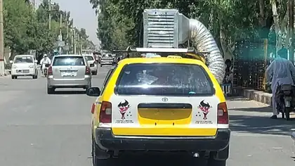 A yellow taxi driving down a street with a full air conditioning duct unit strapped to its roof.