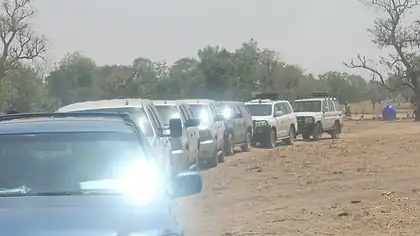 A queue of identical white four wheel drives on a dirt road with headlights on.