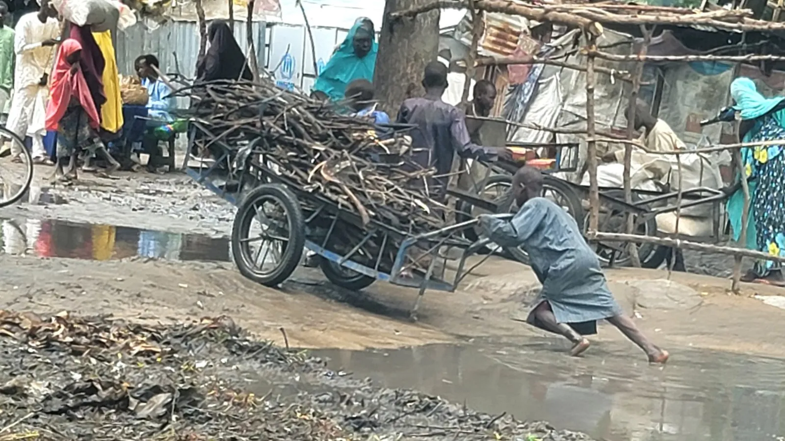 A man hauling an overloaded cart of scrap and firewood through mud by hand.