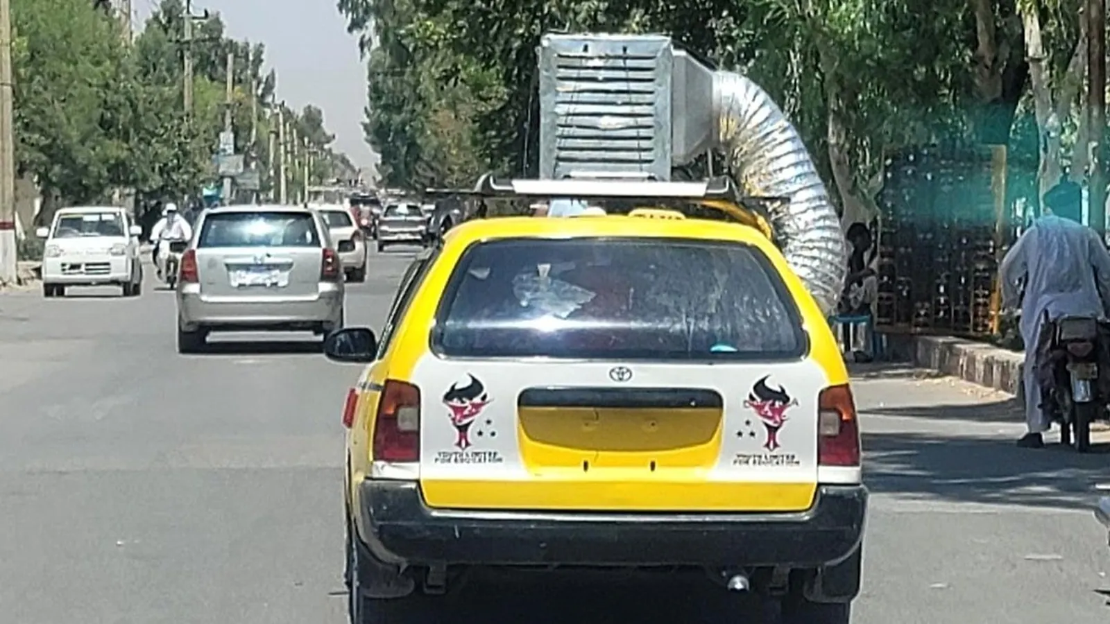 A yellow taxi driving down a street with a full air conditioning duct unit strapped to its roof.