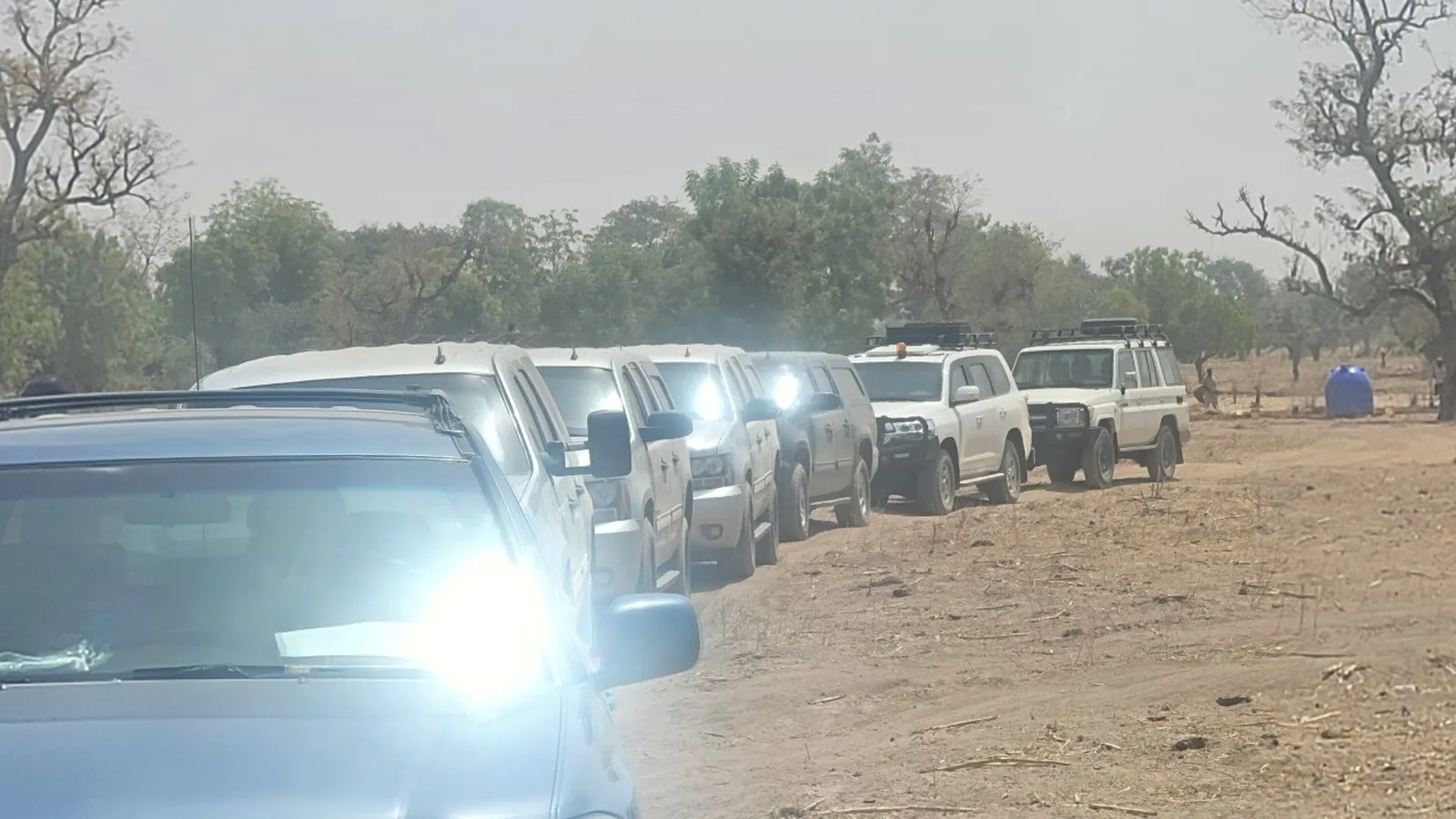 A queue of identical white four wheel drives on a dirt road with headlights on.